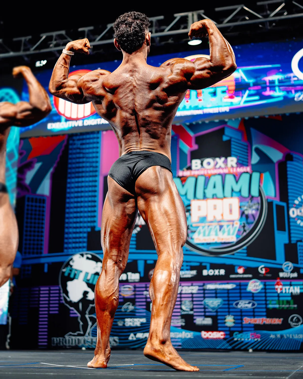 Classic physique trunks in matte black lycra, back view on stage at NPC classic physique competition, regular cut with division-compliant coverage